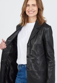 Schwarze Lederjacke mit Knopfverschluss, Reverskragen und Seitentaschen. Offen getragen über einem weißen T-Shirt und blauen Jeans. Glatte Textur.