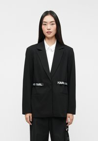 Μαύρο σακάκι με κομψή εφαρμογή, διαθέτοντας δύο μπροστινές τσέπες και λευκά διακοσμητικά στοιχεία. Συνδυασμένο με λευκό πουκάμισο με κολάρο από κάτω.