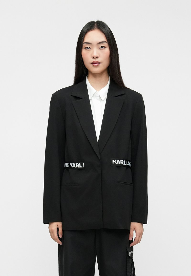 Μαύρο σακάκι με κομψή εφαρμογή, διαθέτοντας δύο μπροστινές τσέπες και λευκά διακοσμητικά στοιχεία. Συνδυασμένο με λευκό πουκάμισο με κολάρο από κάτω.