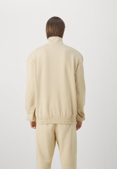 Felpa beige con colletto alto, vestibilità rilassata e vita elasticizzata. Texture morbida con cuciture minime, mostrata dalla parte posteriore.