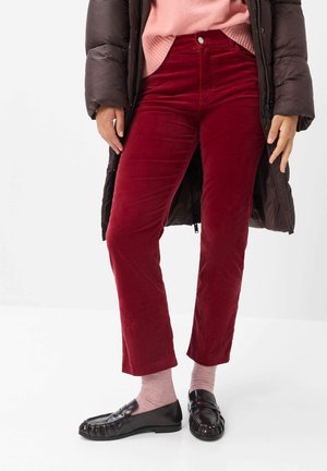 Personne portant un pantalon en velours côtelé bordeaux, des chaussettes roses, des mocassins noirs, un pull rose et un long manteau matelassé marron foncé, debout sur un sol blanc.