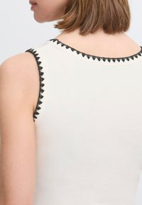 Bezramenný bílý top s černým zigzagovým výšivkovým lemováním kolem výstřihu a otvorů na ruce, zobrazený zezadu. Hladká textura látky.