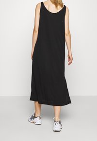 Schwarzes, ärmelloses Maxikleid mit lässiger Passform, rundem Ausschnitt, seitlichen Schlitzen und einer glatten Stofftextur. Kombiniert mit Sportschuhen.