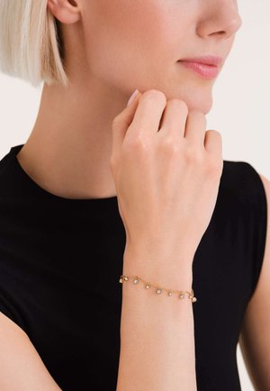 Goldenes Armband mit kleinen, runden, klaren Steinen, die in einer zarten Kette eingefasst sind. Glatte Textur, dezenter Glanz, getragen an einem Handgelenk mit hellem Hautton.