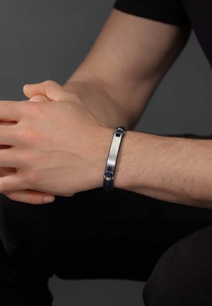 Bracelet en caoutchouc marine avec fermoir en acier inoxydable ; présente un design plat et un motif de maillons texturé. Porté au poignet, les mains jointes.