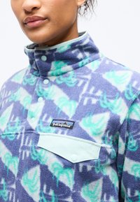 Κοντινό πλάνο ατόμου που φοράει φλις πουλόβερ με μοτίβο σε αποχρώσεις μωβ και μέντας, με κουμπιά τύπου snap και λογότυπο Patagonia στην τσέπη στο στήθος.