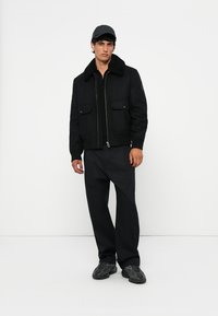 Chaqueta bomber negra con cremallera y cuello de borreguillo texturizado, bolsillos frontales y pantalones negros. Modelo vistiendo una gorra a juego y zapatillas.