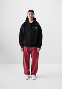 Black zip-up hoodie with green graphic text, kangaroo pocket, and drawstrings, paired with red loose-fit pants and white sneakers.
