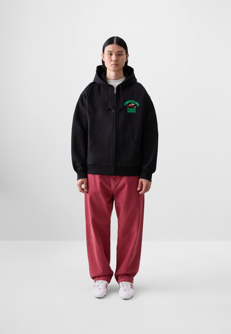 Sweat-shirt noir zippé avec texte graphique vert, poche kangourou et cordons, associé à un pantalon rouge ample et des baskets blanches.