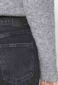 Suéter gris texturizado que cubre parcialmente la cinturilla de unos pantalones vaqueros negros de mezclilla con un bolsillo trasero visible y una etiqueta de cuero de marca.
