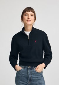 Svart stickad tröja med kort dragkedja, ribbade ärmslut och en liten röd logotyp. Matchad med blekta blå jeans. Mjuk textur, avslappnad passform.
