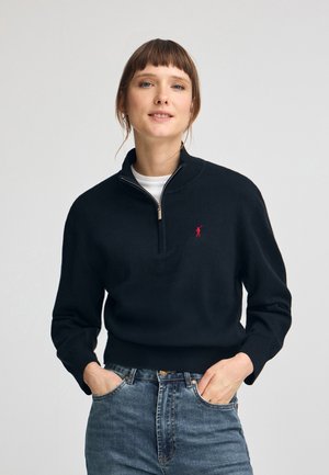 Maglione nero a maglia con una cerniera corta, polsini a costine e un piccolo logo rosso. Abbinato a jeans blu sbiaditi. Tessuto morbido, vestibilità rilassata.