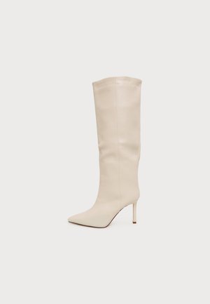 Botte crème montant jusqu’au genou, à bout pointu, talon haut et fin et tige lisse, présentée de profil sur un fond uni.