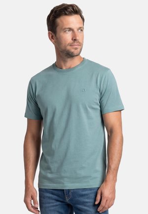 Mann mit kurzen braunen Haaren, der ein hellblaues T-Shirt mit Rundhalsausschnitt und eine blaue Jeans trägt, schaut leicht nach links.