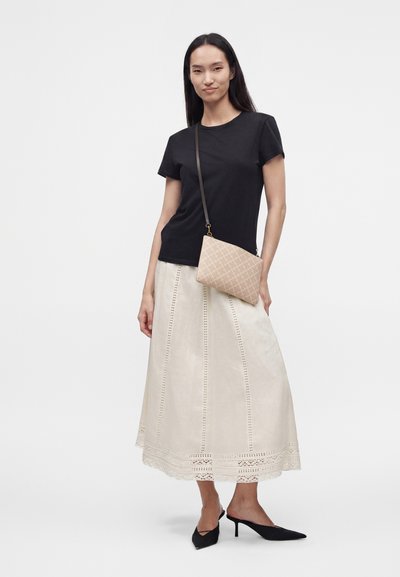 Femme portant un haut noir à manches courtes, une jupe midi beige avec des détails en dentelle, des talons pointus noirs et un sac bandoulière beige, debout sur un fond blanc.