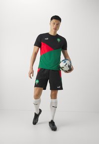 Equipación de fútbol Puma que presenta una camiseta negra con paneles geométricos verdes y rojos, combinada con pantalones cortos negros, calcetines blancos y botas negras.
