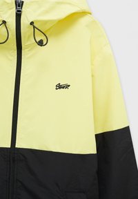 Veste jaune et noire à capuche avec fermeture éclair frontale, cordons de capuche ajustables et petit logo noir sur la poitrine.