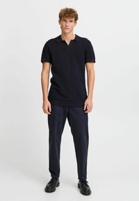 Navy knit polo shirt with short sleeves, v-neck, and a textured finish, paired with dark trousers and black lace-up shoes.