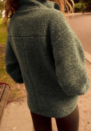 Zielona kurtka z polaru o teksturowanej, miękkiej powierzchni, z wysokim kołnierzem i luźnym krojem. Szczegóły obejmują szew na plecach oraz szerokie rękawy.