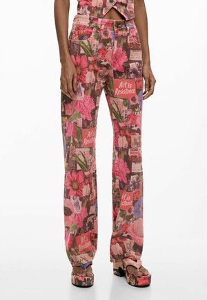 Nő virágmintás foltos nadrágban és hozzá illő platform szandálban, amelyeken "A művészet az ellenállás" felirat látható rózsaszín virágok és zöld levelek között.