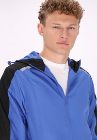 Giacca leggera blu con zip e dettagli neri, dotata di cappuccio, zip frontale e rifiniture riflettenti sulle spalle.