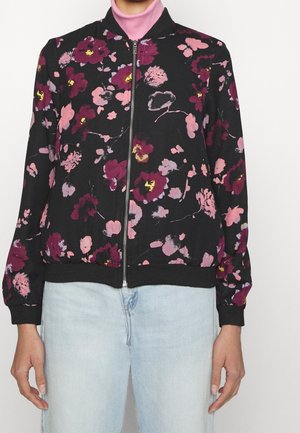 Sort lynlåsjakke med pink og lilla blomsterprint, båret over en pink rullekrave og lyseblå jeans, vist fra skuldre til hofter.