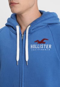 Blå hoodie i mjukt tyg, med dragkedja, vita dragsnören och en röd broderad "Hollister California"-logotyp på bröstet.