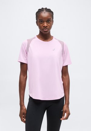 Jeune femme aux cheveux tressés portant un t-shirt de sport rose clair à manches courtes et des leggings noirs, debout devant un fond blanc uni.