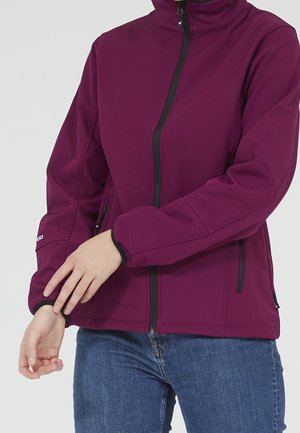 Kobieta poprawiająca rękaw fioletowej kurtki na zamek błyskawiczny w połączeniu z niebieskimi dżinsami na jednolitym tle.