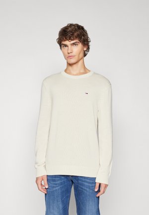 Suéter de punto beige claro con un cuello redondo acanalado, mangas largas y un pequeño parche con el logo en el lado izquierdo del pecho. Llevado con jeans azules.