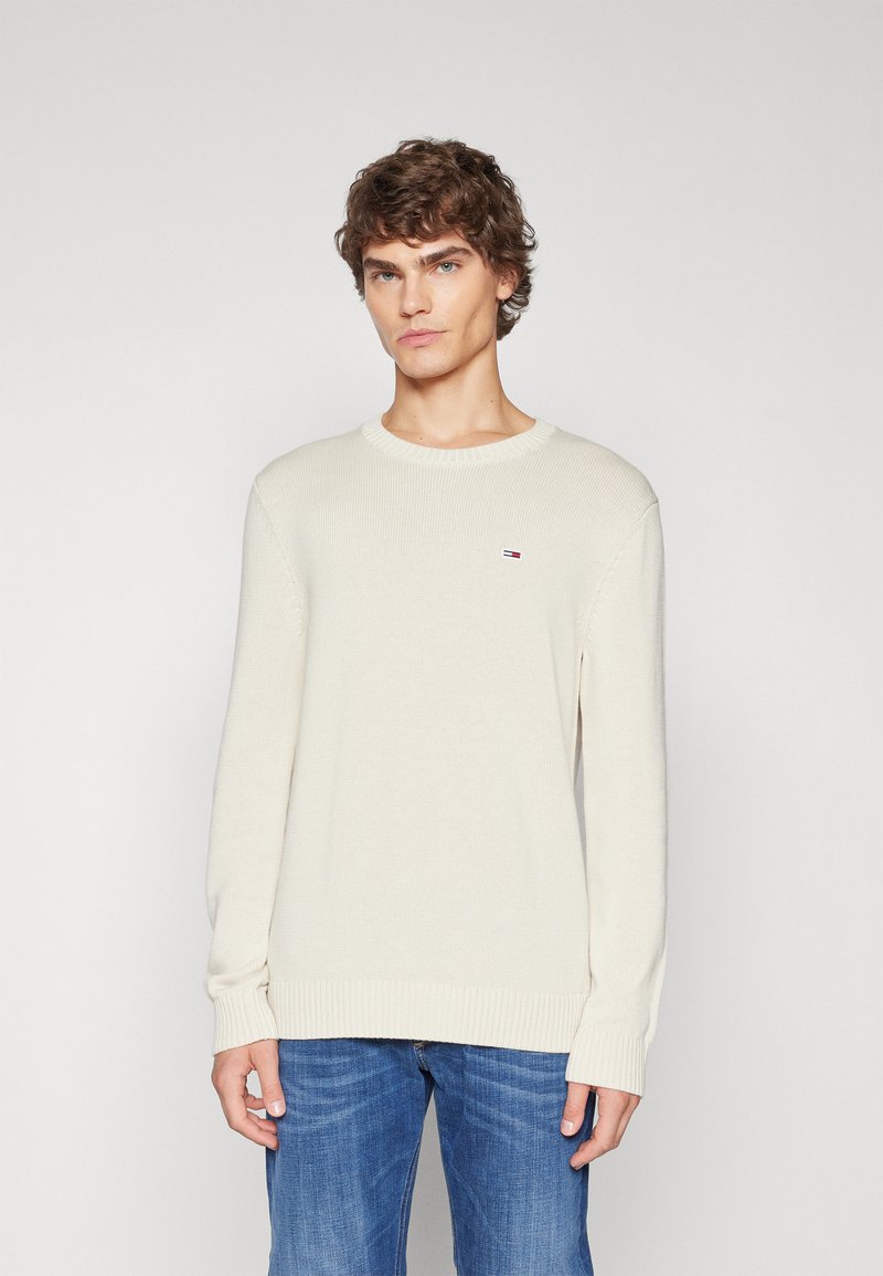 Suéter de punto beige claro con un cuello redondo acanalado, mangas largas y un pequeño parche con el logo en el lado izquierdo del pecho. Llevado con jeans azules.