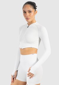 Hvid cropped zip-up top med lange ærmer, ribbet tekstur, parret med højtaljede hvide shorts, glat stof, minimal brandingdetalje.