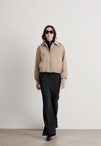 Beige bomber jacket with a zip front, a high collar, and elastic cuffs, paired with black pinstriped wide-leg trousers. Black sunglasses worn.