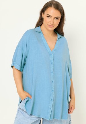 Frau trägt ein locker sitzendes hellblaues Hemd mit kurzen, hochgekrempelten Ärmeln und hellblaue Jeans, posiert mit den Händen in den Taschen.