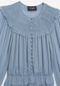 Blusa azzurra chiara con un design a pieghe, colletto arricciato e bottoni decorativi sul davanti. Tessuto morbido e strutturato con vita arricciata.