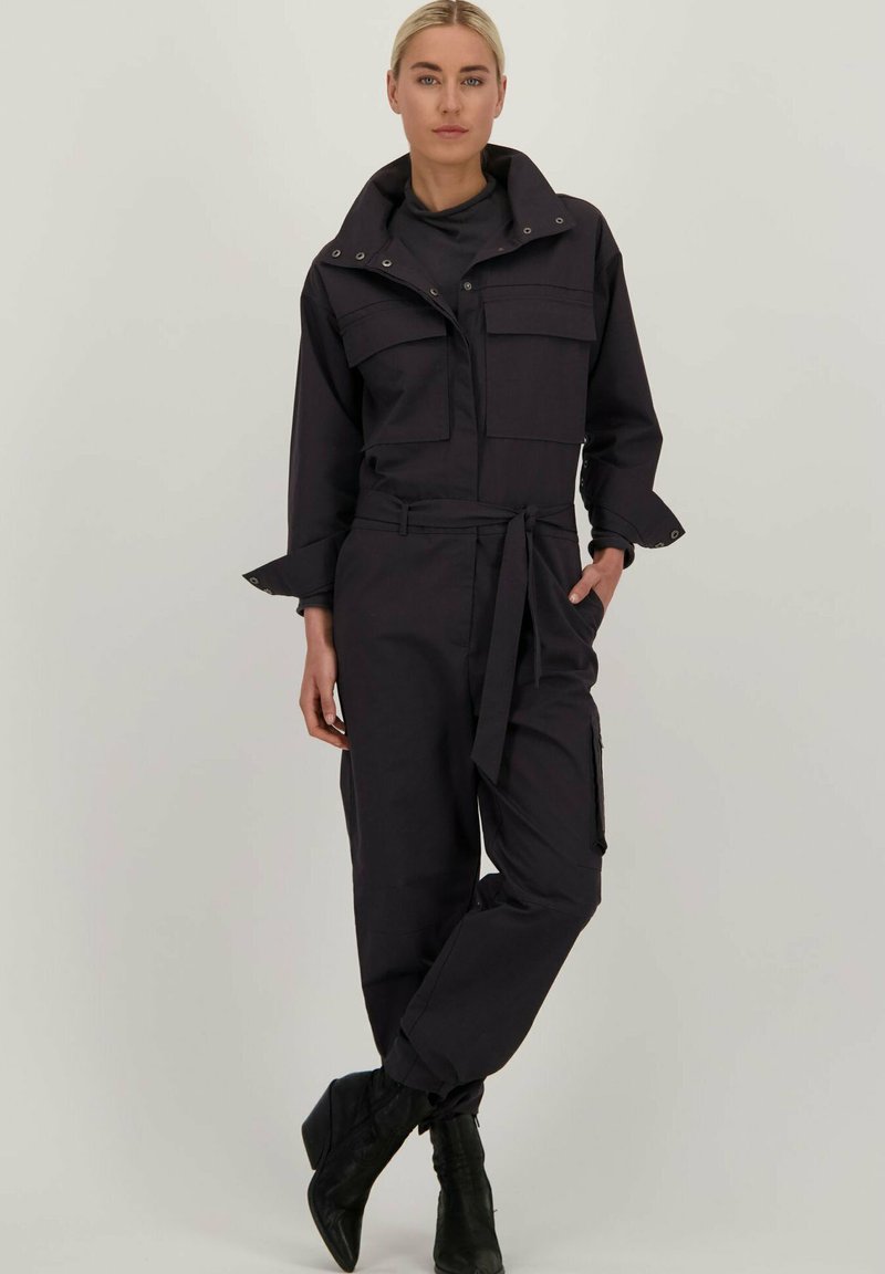 Zwarte jumpsuit met een hoge kraag, voorzakken en een ceintuur in de taille. De chenille textuur en taps toelopende pijpen versterken het gestructureerde ontwerp.