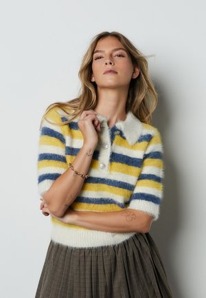 Donna con capelli biondi lunghi che indossa un maglione corto a righe gialle, bianche e blu, soffice, con bottoni di strass e una gonna a quadri.