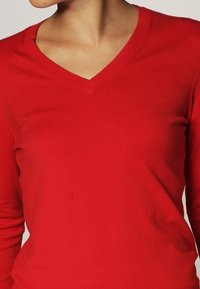 Pull over rouge en V, fait de tissu côtes. Manches longues, design ajusté, avec un décolleté épuré et sans motifs ni accents visibles.