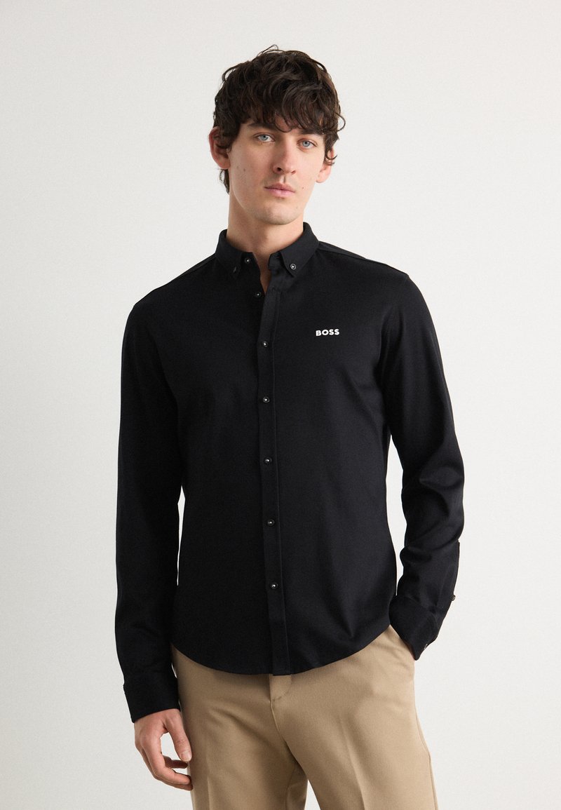 Zwarte blouse met lange mouwen en kraag, voorzien van een klein wit "BOSS" logo op de borst, gecombineerd met lichte kaki broeken.