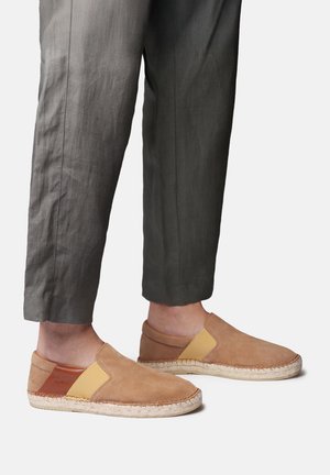 Person trägt braune Slip-On-Suede-Espadrilles mit gelben elastischen Einsätzen und dunkelgraue, gekürzte Hosen vor weißem Hintergrund.