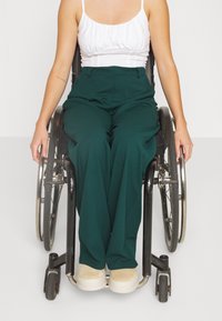 Person sitzt im Rollstuhl, trägt ein weißes ärmelloses Oberteil, grüne Hosen und beige Schuhe, die Hände ruhen auf den Rollstuhlrädern.
