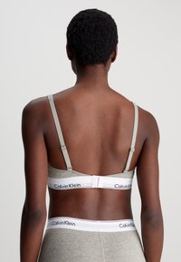 Grå stoff-bh med justerbare stropper og en hvit bånd med Calvin Klein-logoen. Ingen synlige mønstre eller ekstra maskinvare.