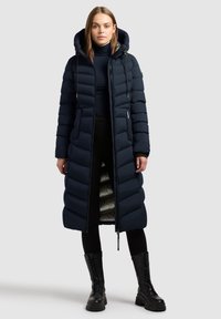Lang navy pufferjakke med hætte, der har lodret quiltet design, lynlås lukning og en reflekterende indvendig foring. Båret over sorte bukser og støvler.