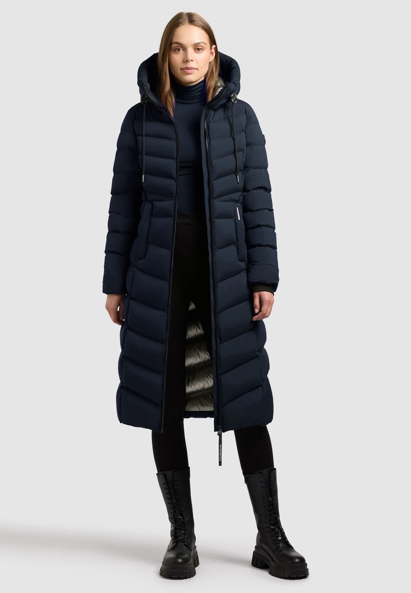 Lang navy pufferjakke med hætte, der har lodret quiltet design, lynlås lukning og en reflekterende indvendig foring. Båret over sorte bukser og støvler.