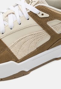 Sneaker mit einem Obermaterial aus tanfarbigem Wildleder und cremefarbenem Leder, weißen Schnürsenkeln und einer gepolsterten weißen Sohle mit ausgeprägter Textur und Designelementen.