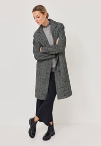 Palton lung texturat de culoare gri, cu guler, purtat peste un pulover gri deschis și pantaloni negri cu croi larg. Botine negre până la glezne completează ținuta.