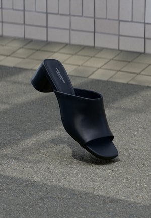 Mule en cuir noir à bout ouvert avec talon bloc, positionnée sur l'asphalte près d'une surface carrelée en arrière-plan.