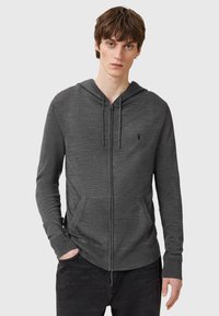 Grå zip-up hoodie tillverkad av mjukt material med en framficka, dragsko i huvan och subtil logotypdetalj på bröstet.