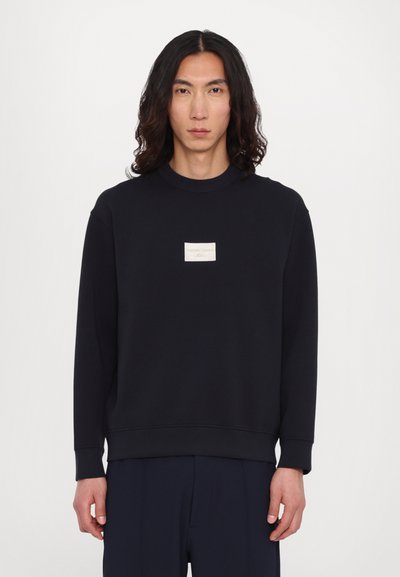 Sweatshirt de un bleu închis, fabricat dintr-un material moale, având un mic patch rectangular cu logo crem pe piept și manșete și tiv cu nervuri.