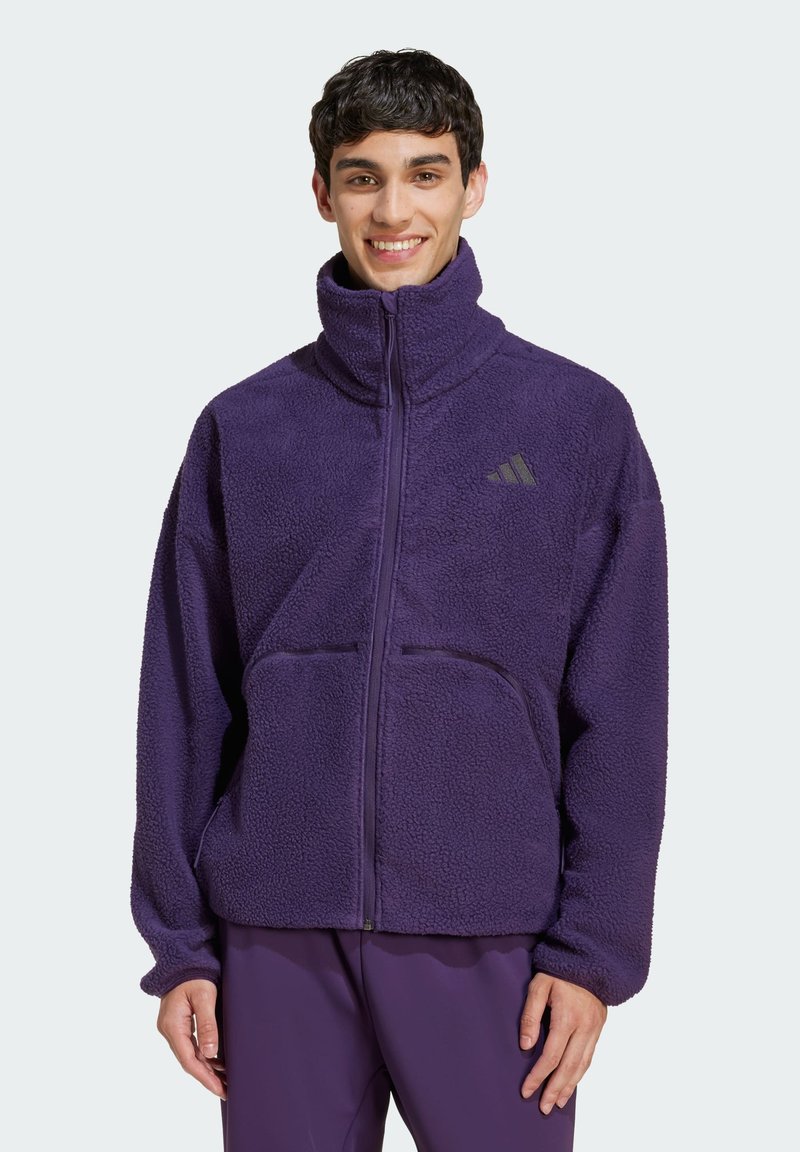 Giacca in pile viola con collo alto, zip frontale e due tasche anteriori. Presenta un logo sul lato sinistro del petto. Materiale morbido e strutturato.